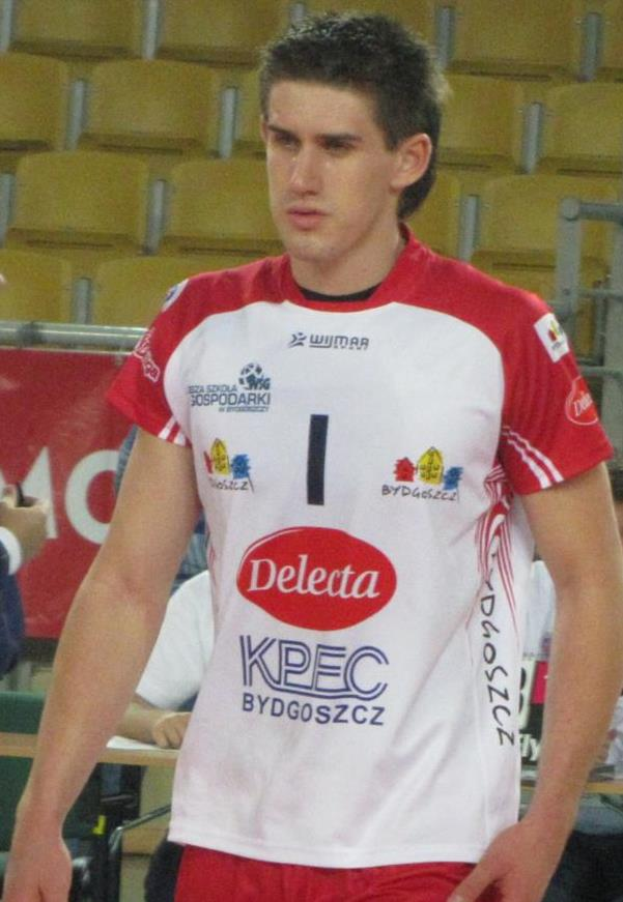Ein Mann in rot-weißer Uniform steht auf einem Handballfeld, umgeben von Zuschauern, mit einem roten Banner im Hintergrund, auf dem 'Krasnoyarsk vs Bydgoszcz' steht.
