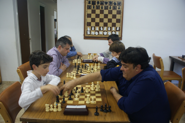 Ein Tisch mit drei Schachbrettern und zahlreichen Schachfiguren in der Mitte, mit drei Menschen, die rechts und links Schach spielen, und drei Menschen, die auf Stühlen sitzen. Eine Wand mit einem Schachbrettplakat in der Mitte, mit einem Tisch und einem Stuhl rechts und einer Tür links.