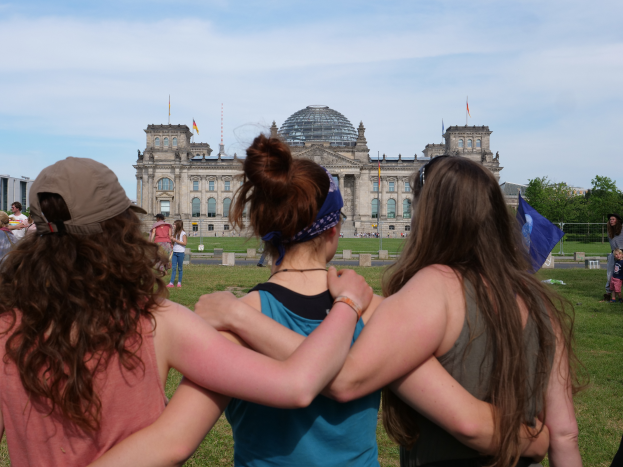 Drei Frauen in verschiedenenfarbigen Kleidern stehen vor dem Reichstaggebäude in Berlin, Deutschland, mit Menschen, Fahnen, Bäumen, Wolken und Himmel im Hintergrund.
