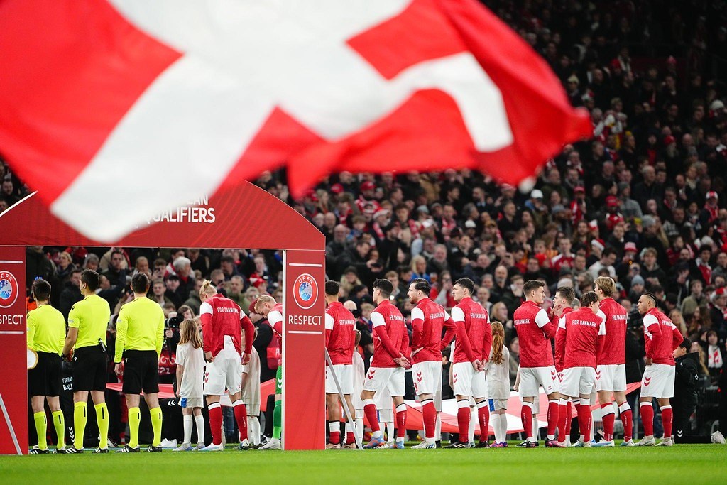 Gruppe von Menschen auf einem Fußballfeld mit einer roten und weißen Flagge im Vordergrund, einem Bogen mit der Aufschrift "Bayern München vs. Bayern München Wetten & Vorschau" im Hintergrund und einer großen Stadionmenge.