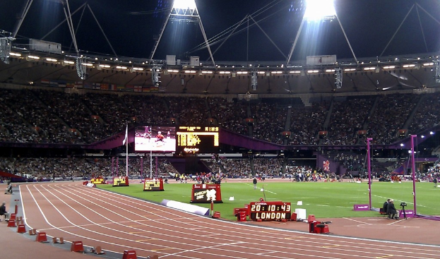Ein Stadion mit einem grasbedeckten Feld, Zuschauern im Pavillon und Lichtern oben, unter einem dunklen Himmel, mit einem Stoppuhr, auf dem "London" steht.