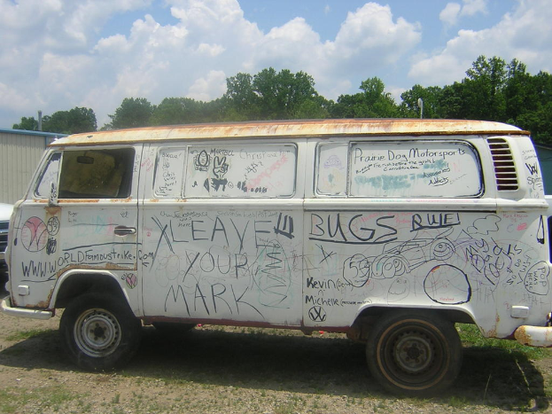 Ein beschädigtes Auto mit Malereien und Zitaten darauf, das vor einem bewaldeten Gebiet geparkt ist.