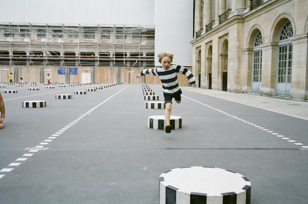 Ein Kind läuft auf einem schwarz-weißen Beton-Spielplatz mit kleinen Stufen, umgeben von einem Schulgebäude, mit drei anderen Kindern im Hintergrund.