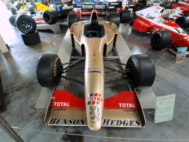 Rennwagen in einem Motorsportmuseum ausgestellt, getrennt durch einen Metallzaun von Zuschauern, mit informativem Text im Hintergrund.