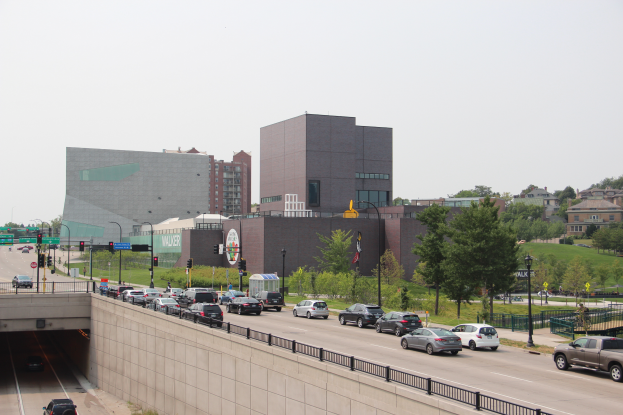 Eine Stadtstraße mit Autos, eine Brücke mit Geländern, Laternen, Verkehrszeichen, Bäumen, Gebäuden mit Fenstern und einem Himmel im Hintergrund.