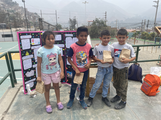 Gruppe von Kindern auf einem Basketballplatz mit Papierbeuteln in der Hand, mit Vorräten auf dem Boden und einer Tafel mit Text links daneben, vor dem Hintergrund von Zäunen, Pfosten, Straßenlaternen, Bäumen, Gebäuden, Hügeln und Himmel.