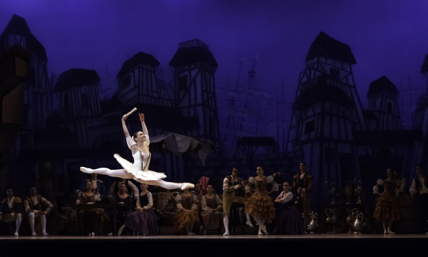 Eine Ballerina in einem weißen Tutu und Spitzenschuhen tanzt auf einer Bühne mit sitzenden und stehenden Zuschauern, während ein Schlossgemälde im Hintergrund sichtbar ist.