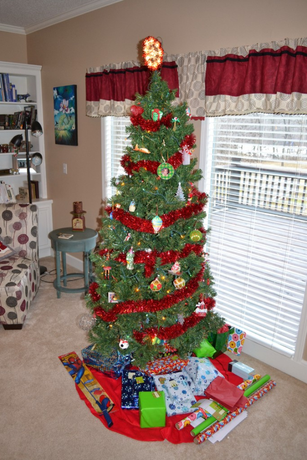 Ein Weihnachtsbaum mit Geschenken davor, eine Couch, ein Tisch, ein Bücherregal, eine Stehlampe und ein Bild an der Wand, alles in einem Raum mit Fenstern und Vorhängen.