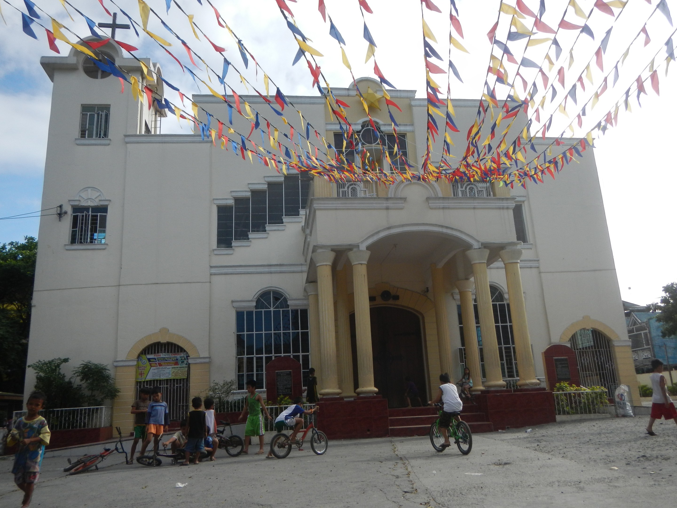 Eine Gruppe von Menschen, die Fahrräder vor einer weißen Kirche mit architektonischen Details fahren, umgeben von Grün, einem Banner, Bäumen und bunten Fahnen vor einem bewölkten Himmel.