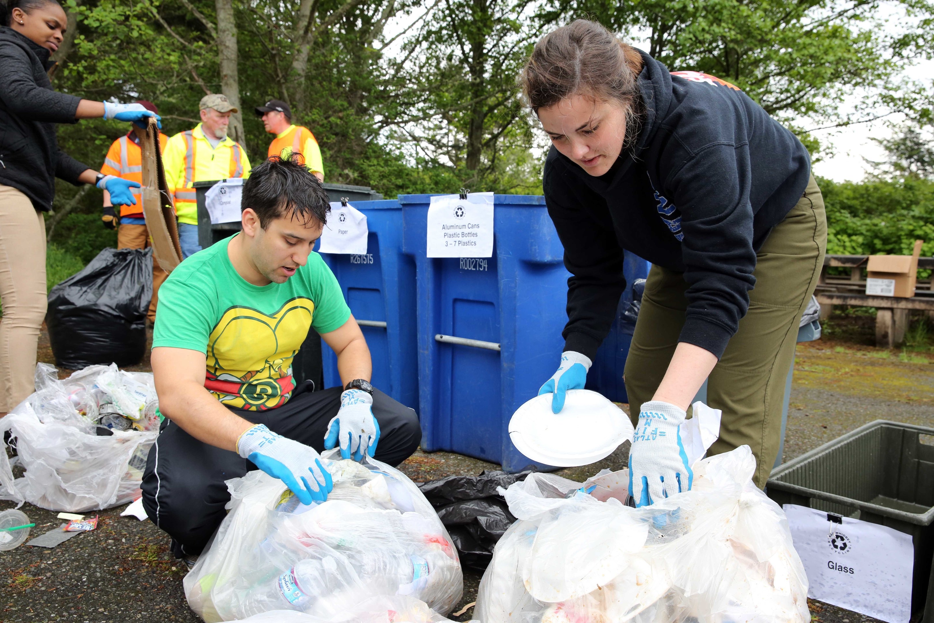 Eine Gruppe von Menschen mit Handschuhen sammelt Müll in einem Park, mit Plastikabfällen, Flaschen und anderem Müll auf dem Boden in der Nähe eines Mülleimers und einer hölzernen Bank, vor einem Hintergrund von Bäumen und einem klaren Himmel.