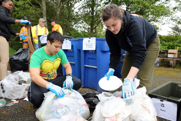 Eine Gruppe von Menschen mit Handschuhen sammelt Müll in einem Park, mit Plastikabfällen, Flaschen und anderem Müll auf dem Boden in der Nähe eines Mülleimers und einer hölzernen Bank, vor einem Hintergrund von Bäumen und einem klaren Himmel.