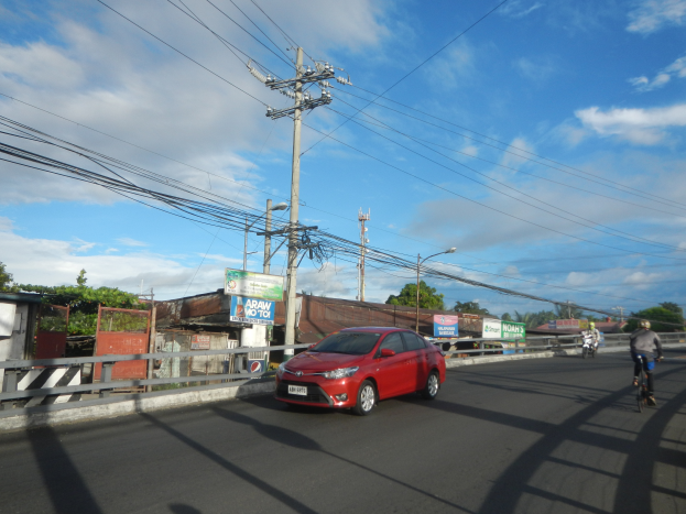 Ein rotes Auto fährt auf einer Straße mit Strommasten und -kabeln entlang, mit ein paar Radfahrern in der Nähe, Gebäuden, Bäumen und einem klaren blauen Himmel im Hintergrund.