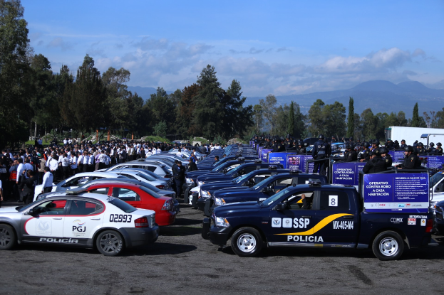Große Anzahl von Polizeiwagen auf einem Parkplatz mit Menschen in der Nähe, Schilder mit Text, Bäume, Hügel und bewölkter Himmel im Hintergrund.