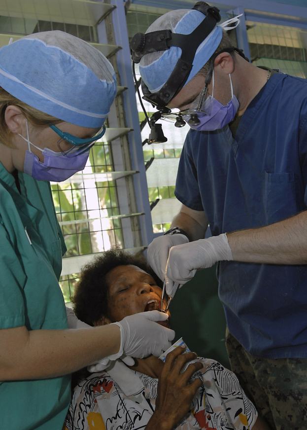 Zwei maskierte und handschuhgetragene Chirurgen führen eine Gesichtsoperation an einer Frau durch, die auf einem Bett liegt, mit einem Fenster und Bäumen im Hintergrund.