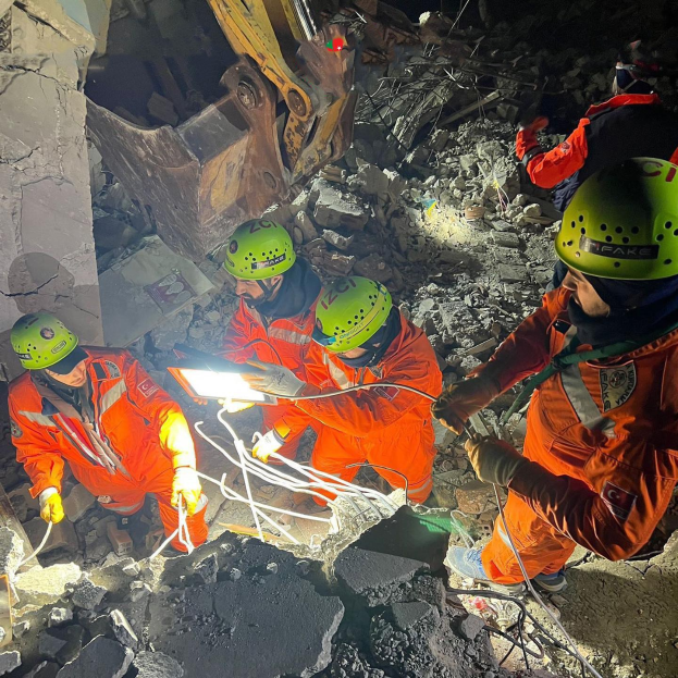 Gruppe von Menschen in orangefarbenen Uniformen und grünen Helmen bei der Arbeit an Schutt von einem eingestürzten Gebäude mit einem Kran im Hintergrund, hölden Rettungsgeräte.