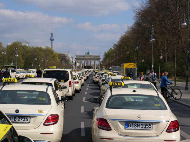 Eine lange Reihe von Taxis, die an einer belebten Straße in Berlin, Deutschland, geparkt sind, mit Fahrradfahrern und Fußgängern auf dem Gehweg, flankiert von Bäumen und Laternenpfählen und Gebäuden, einem Bogen und einem Turm im Hintergrund unter einem bewölkten Himmel.