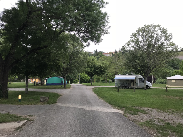 Campingplatz mit einem parkenden Auto neben einem Baum, grasbewachsenem Boden, verstreuten Zelten und Bänken, Bäumen und Gebäuden im Hintergrund unter einem klaren blauen Himmel.